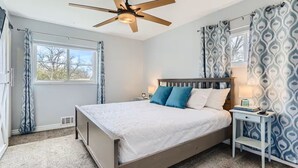 Ein Schlafzimmer mit einem Bett, einem Nachttisch, einem Deckenventilator und gemusterten Vorhängen.