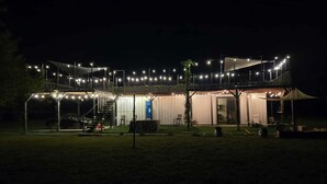 A two-story building with a covered outdoor area, a staircase, and string lights. - Cozy-Chic Container Tiny Home + Treehouse Deck (Wagoner)