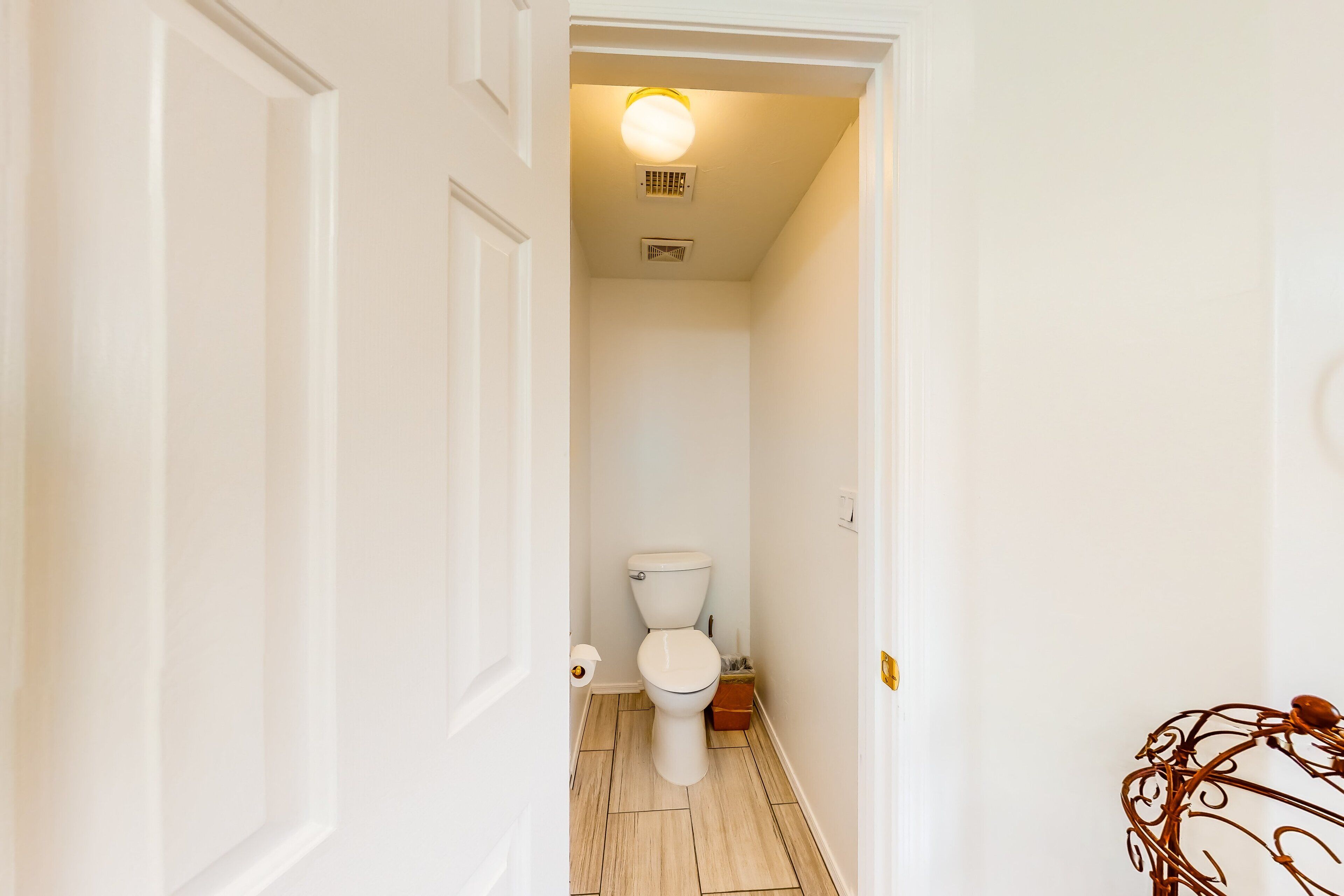 Un couloir étroit mène à une salle de bain équipée de toilettes, d’une petite poubelle et d’un luminaire au plafond.