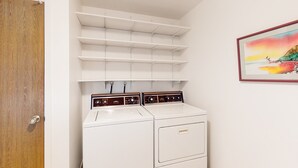 A laundry room with a white washer and dryer, shelves above, and a painting on the wall. - Snowbird-friendly golf-front home with pool, hot tub, grill, smart TVs, W/D, AC (Mesa)