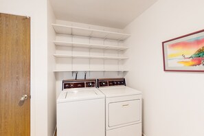 A laundry room with a white washer and dryer, shelves above, and a painting on the wall. - Snowbird-friendly golf-front home with pool, hot tub, grill, smart TVs, W/D, AC (Mesa)
