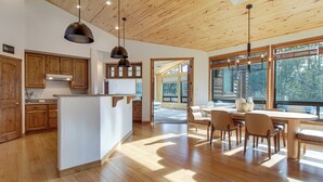 Ett modernt kök med träskåp, en köksö, hängande lampor och stora fönster som ger utsikt över omgivningen.