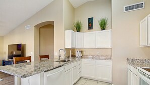 Una cocina con gabinetes blancos, encimeras de granito y un horno empotrado. Se ve un área de comedor con una mesa y sillas al fondo.