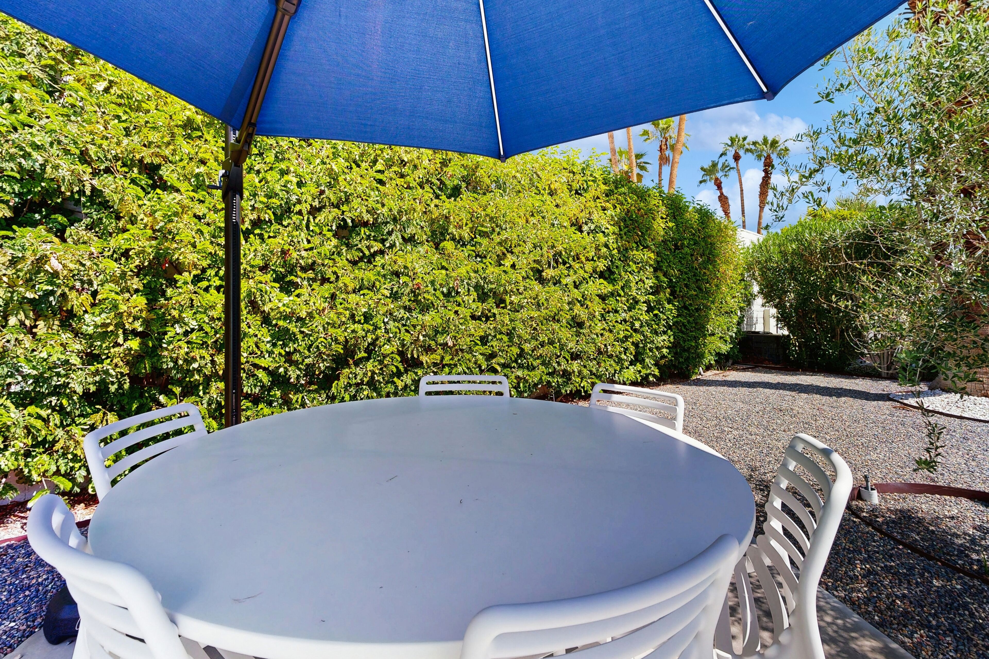 Een ronde tafel met witte stoelen onder een blauwe parasol, omringd door weelderig groen.