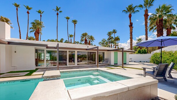 A modern outdoor pool area with a clear blue pool, surrounded by palm trees and a covered seating area with chairs. - Poolside Paradise in the Desert – Your Perfect Palm Springs Escape Awaits. (Palm Springs)