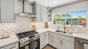 A modern kitchen with white cabinets, a stainless steel stove, and a window with a view of greenery. - Designer dream with fireplace, game room, private pool, outdoor kitchen, firepit (Palm Springs)