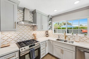 A modern kitchen with white cabinets, a stainless steel stove, and a window with a view of greenery. - Designer dream with fireplace, game room, private pool, outdoor kitchen, firepit (Palm Springs)
