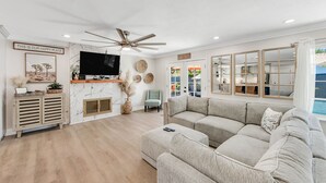 A spacious living room with a large sectional sofa, a flat-screen TV, a ceiling fan, and a view of the outdoors through large windows. - Designer dream with fireplace, game room, private pool, outdoor kitchen, firepit (Palm Springs)