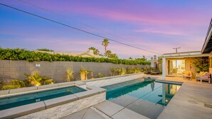 A swimming pool with a stone border, a patio area with seating, and a view of a residential building in the background. - Designer dream with fireplace, game room, private pool, outdoor kitchen, firepit (Palm Springs)