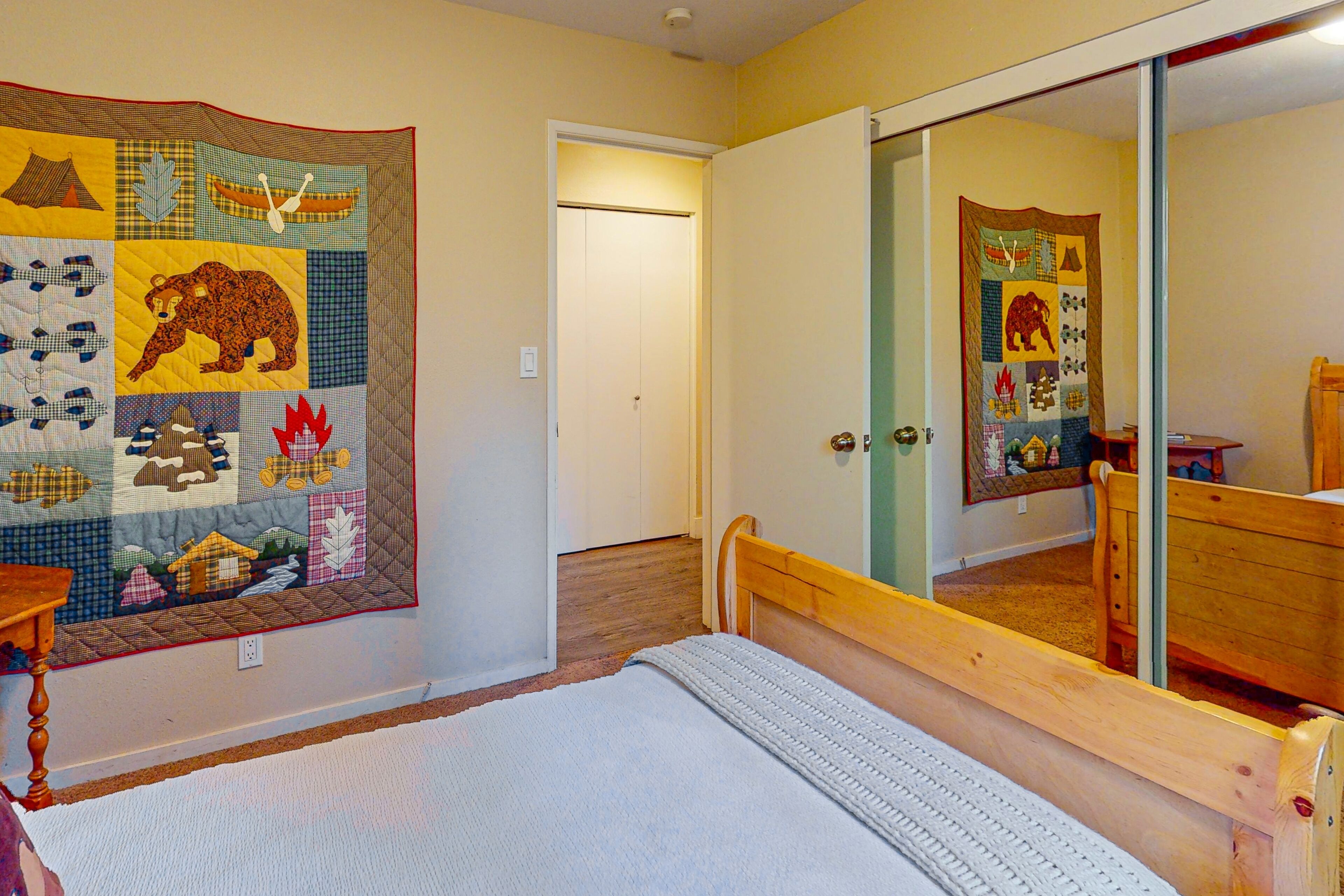 A bedroom with a quilted wall hanging, a wooden bed, and a mirror reflecting the room.