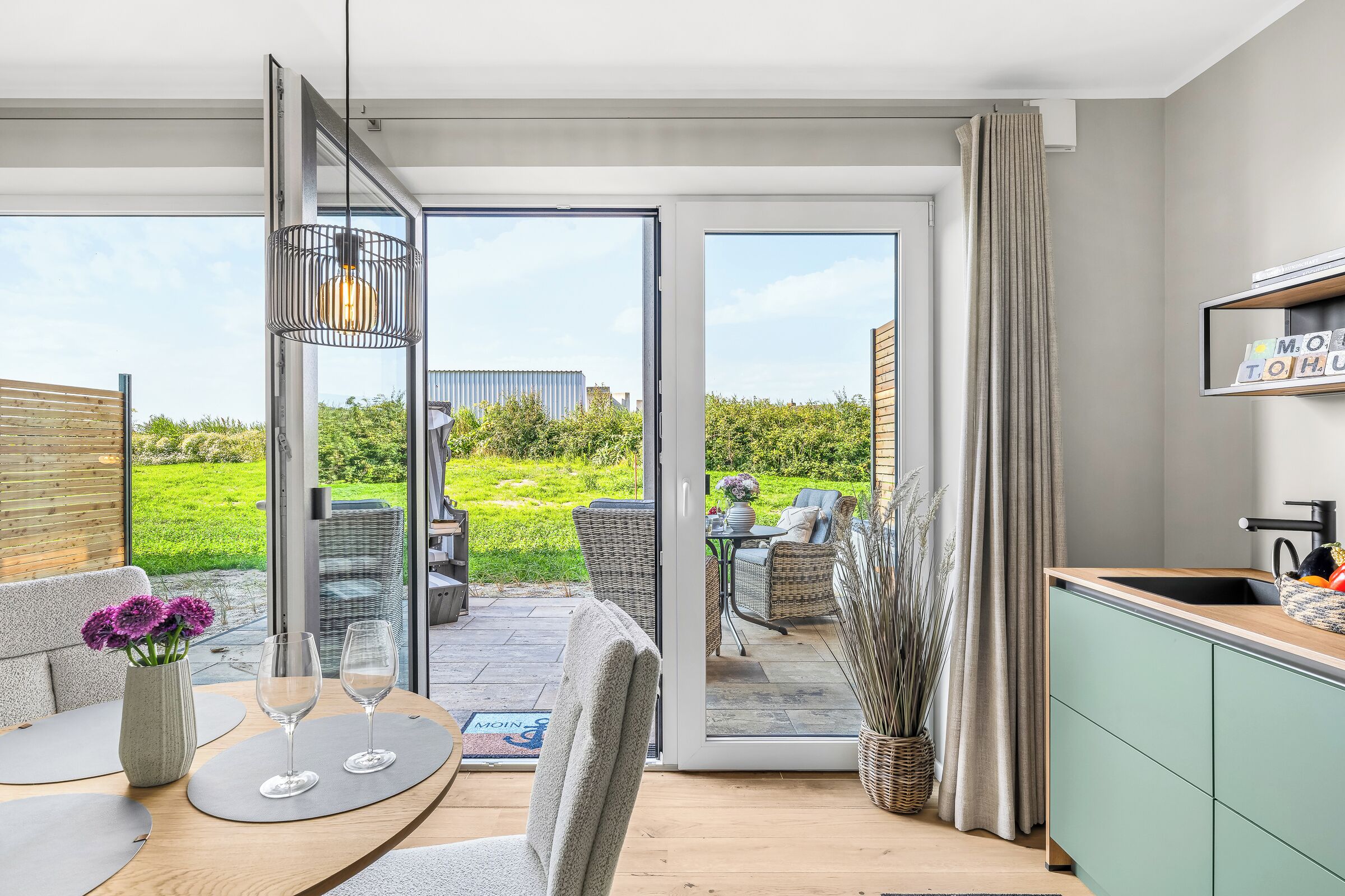 Ein modernes Wohnzimmer mit Glastür zum Außenbereich, ein Essbereich mit Tisch und Stühlen sowie eine Küche mit Spüle und Schränken.