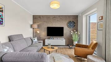 Ein modernes Wohnzimmer mit einem grauen Sofa, einem braunen Sessel, einem Couchtisch, einem Flachbildfernseher, einer Uhr und einem Fenster mit Vorhängen.