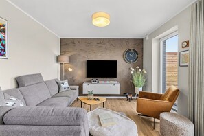 Ein modernes Wohnzimmer mit einem grauen Sofa, einem braunen Sessel, einem Couchtisch, einem Flachbildfernseher, einer Uhr und einem Fenster mit Vorhängen.