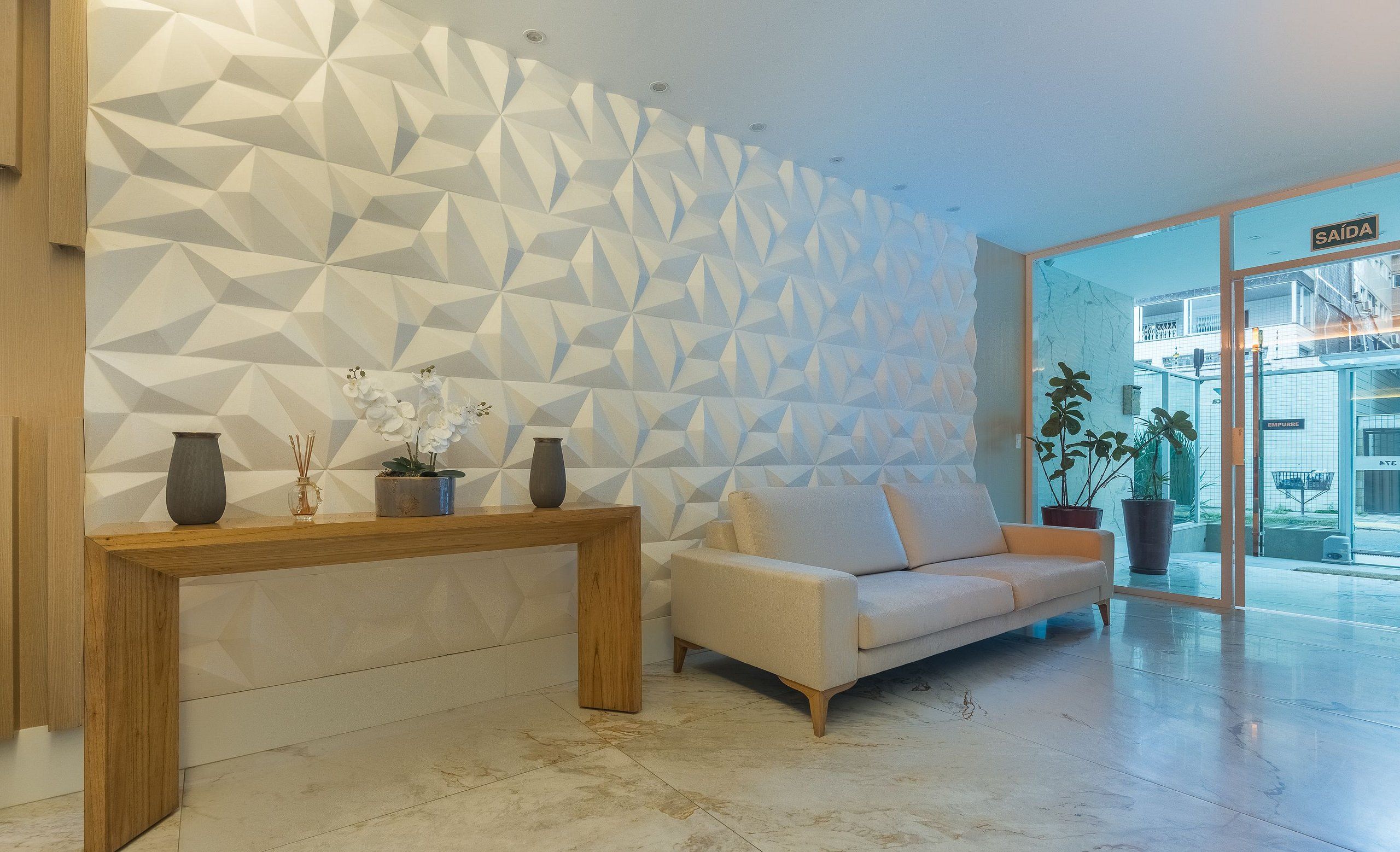 Un interior moderno con una pared de diseño geométrico, una consola de madera, un sofá color beige y plantas en macetas.