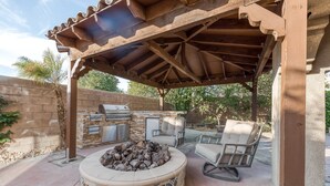 En uteplats med en eldstad i sten, en grill och två bekväma fåtöljer under en träpergola.