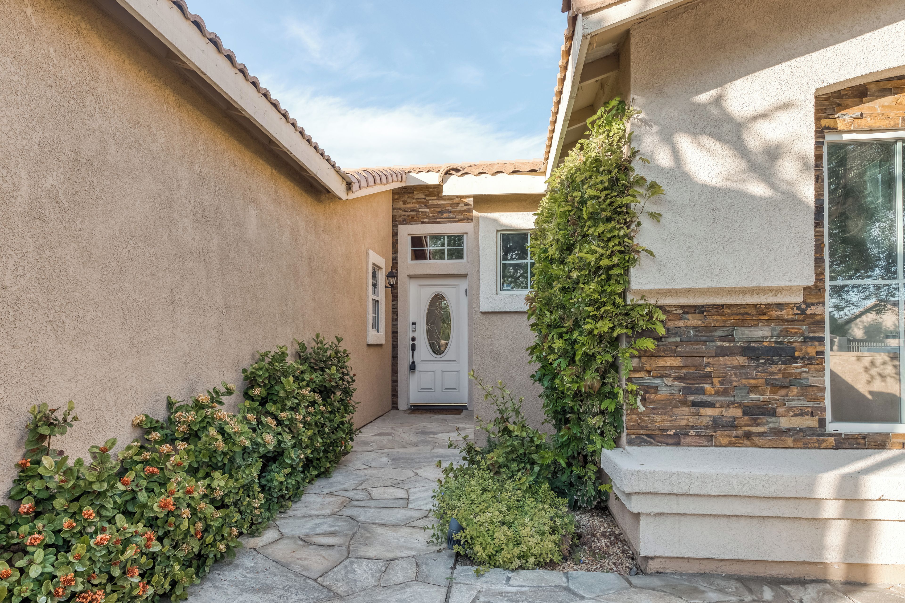 A house with a stone pathway leading to a white door, surrounded by greenery and a stone wall.