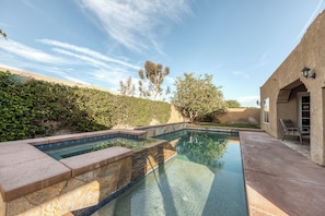 A swimming pool with a stone border, a patio area with a chair, and a building with a light fixture. - Beautiful & spacious home with private pool, spa, and basketball hoop (Indio)