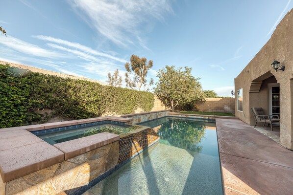 A swimming pool with a stone border, a patio area with a chair, and a building with a light fixture. - Beautiful & spacious home with private pool, spa, and basketball hoop (Indio)
