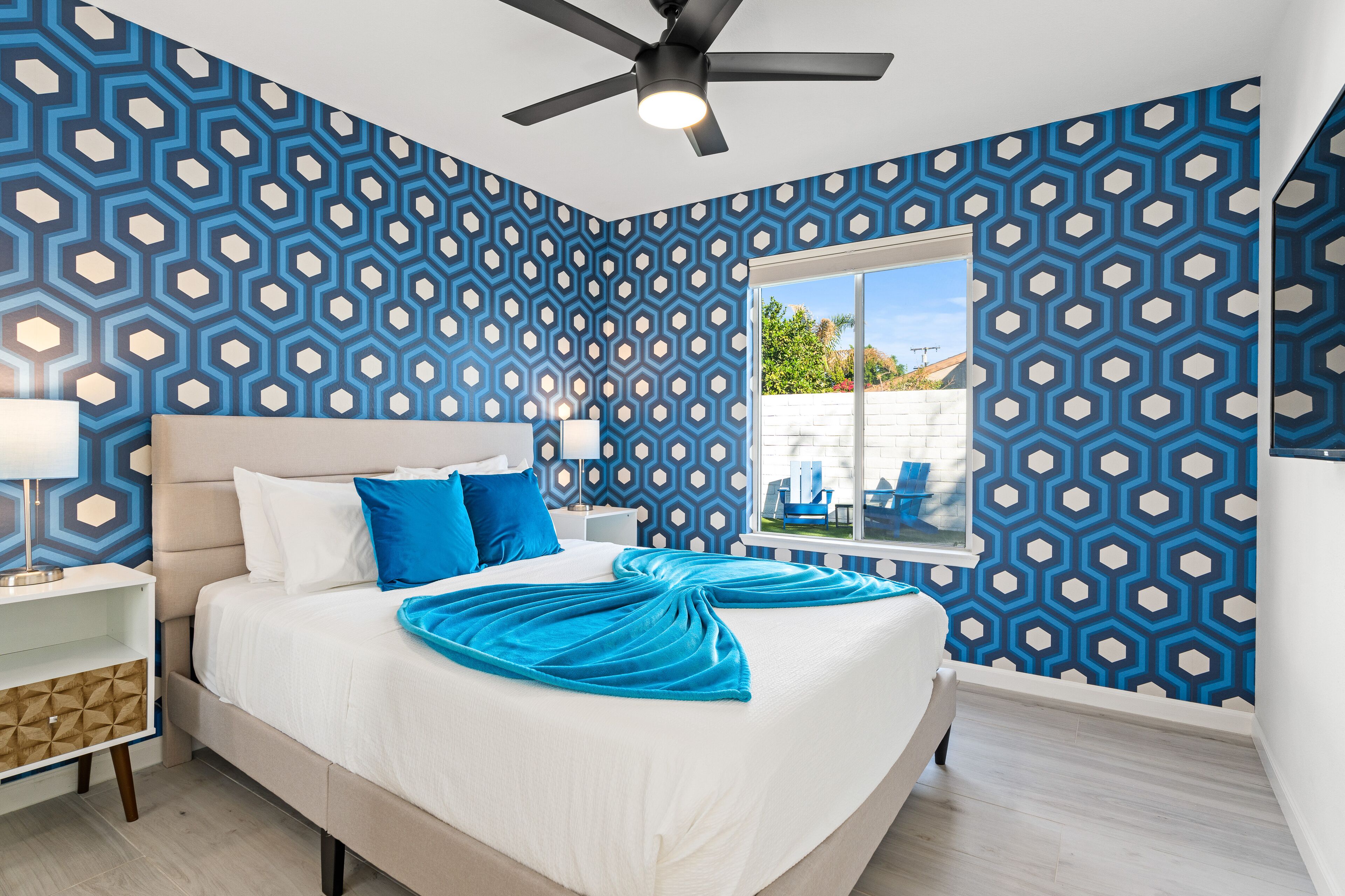 Quarto com cama, mesa de cabeceira, ventilador de teto e janela com vista para o exterior.