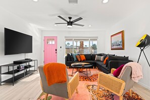 A modern living room with a black entertainment unit, a pink door, a black sofa with colorful cushions, and a large window with a view of palm trees. - Amazing dog-friendly home with enclosed yard, firepit, pool, & washer/dryer (Palm Springs)
