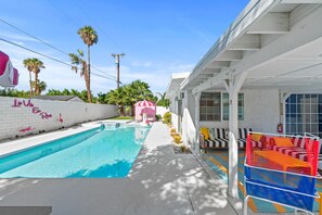 A swimming pool with a white tiled wall and a pink flamingo decoration. There is a white building with a garage door and a colorful outdoor seating area. - Amazing dog-friendly home with enclosed yard, firepit, pool, & washer/dryer (Palm Springs)