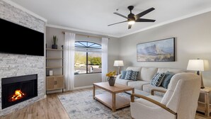 A modern living room with a fireplace, a flat-screen TV, a ceiling fan, a sofa, a coffee table, and a painting on the wall. - Scottsdale Bliss – Stylish Long Term, Comfort, and Mountain Views Await! (Scottsdale)