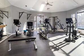 Ein Fitnessraum mit verschiedenen Trainingsgeräten, einem Deckenventilator und großen Fenstern, die natürliches Licht hereinlassen.