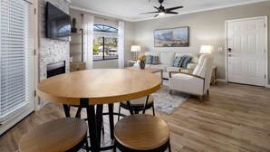 A modern living room with a round wooden table, two chairs, a sofa, and a TV mounted on a stone wall. - Scottsdale Bliss – Stylish Long Term, Comfort, and Mountain Views Await! (Scottsdale)