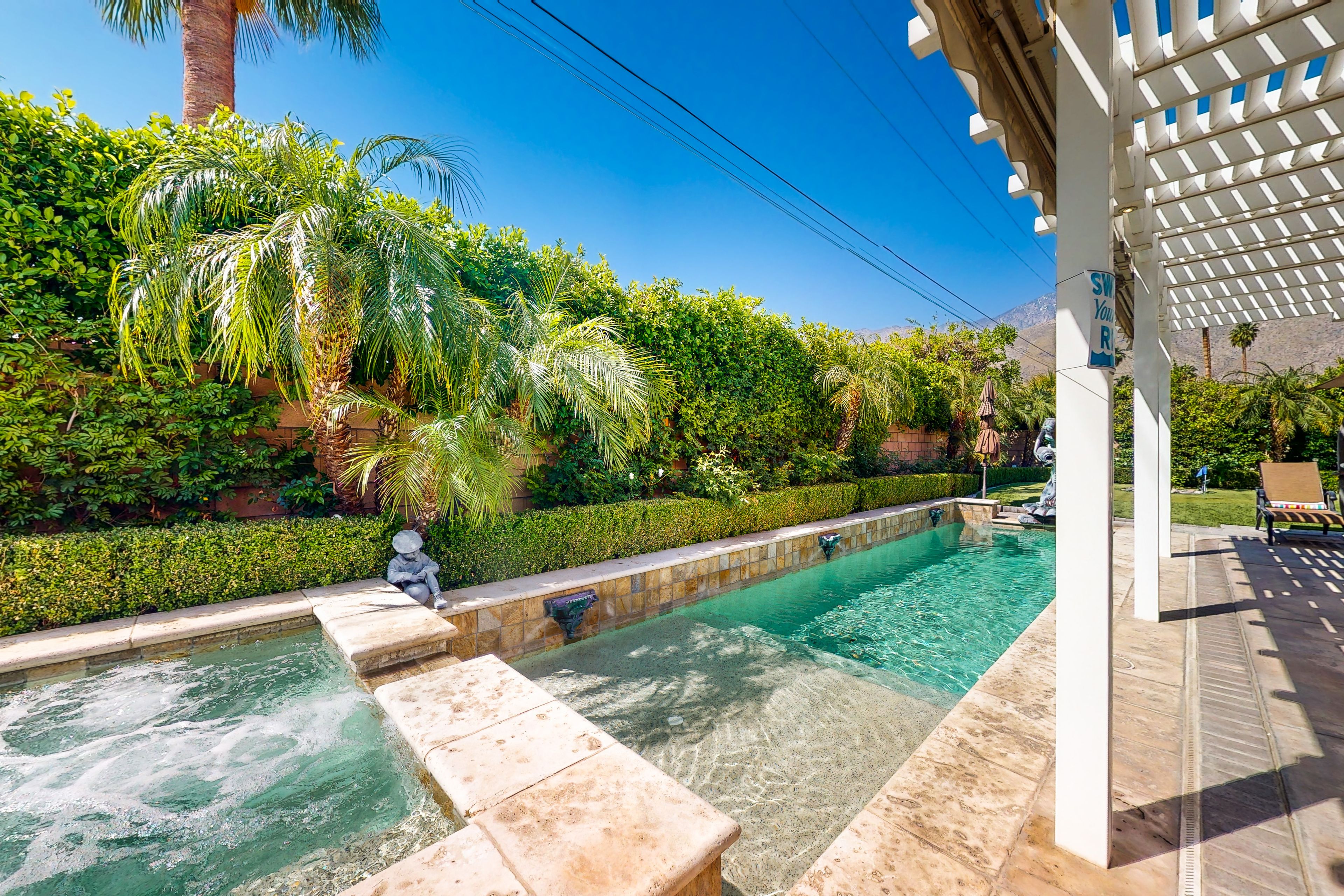 Una piscina con borde de piedra, rodeada de abundante vegetación y una estructura de pérgola.