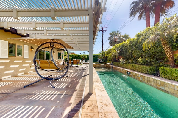 A pool area with a covered seating area, a clear pool, and surrounding greenery. - Elegant home with private pool, spa, putting green, firepit & gorgeous patio (Palm Springs)