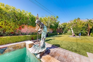 A statue by a pool with a fountain, surrounded by a well-maintained garden and a clear sky. - Elegant home with private pool, spa, putting green, firepit & gorgeous patio (Palm Springs)