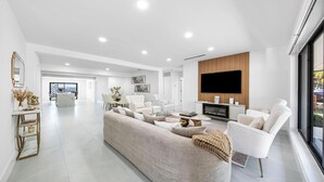 Un salon spacieux avec un grand canapé beige, des fauteuils blancs et un téléviseur à écran plat fixé sur un mur recouvert de panneaux en bois.