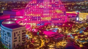 Un hôtel dont la façade, en forme de guitare, est illuminée de lumières roses et rouges, et qui est entouré d’un espace paysager agrémenté de piscines et de palmiers.