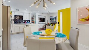 Une cuisine moderne avec des meubles blancs, un coin repas doté d’une table jaune et un luminaire au plafond au design original.