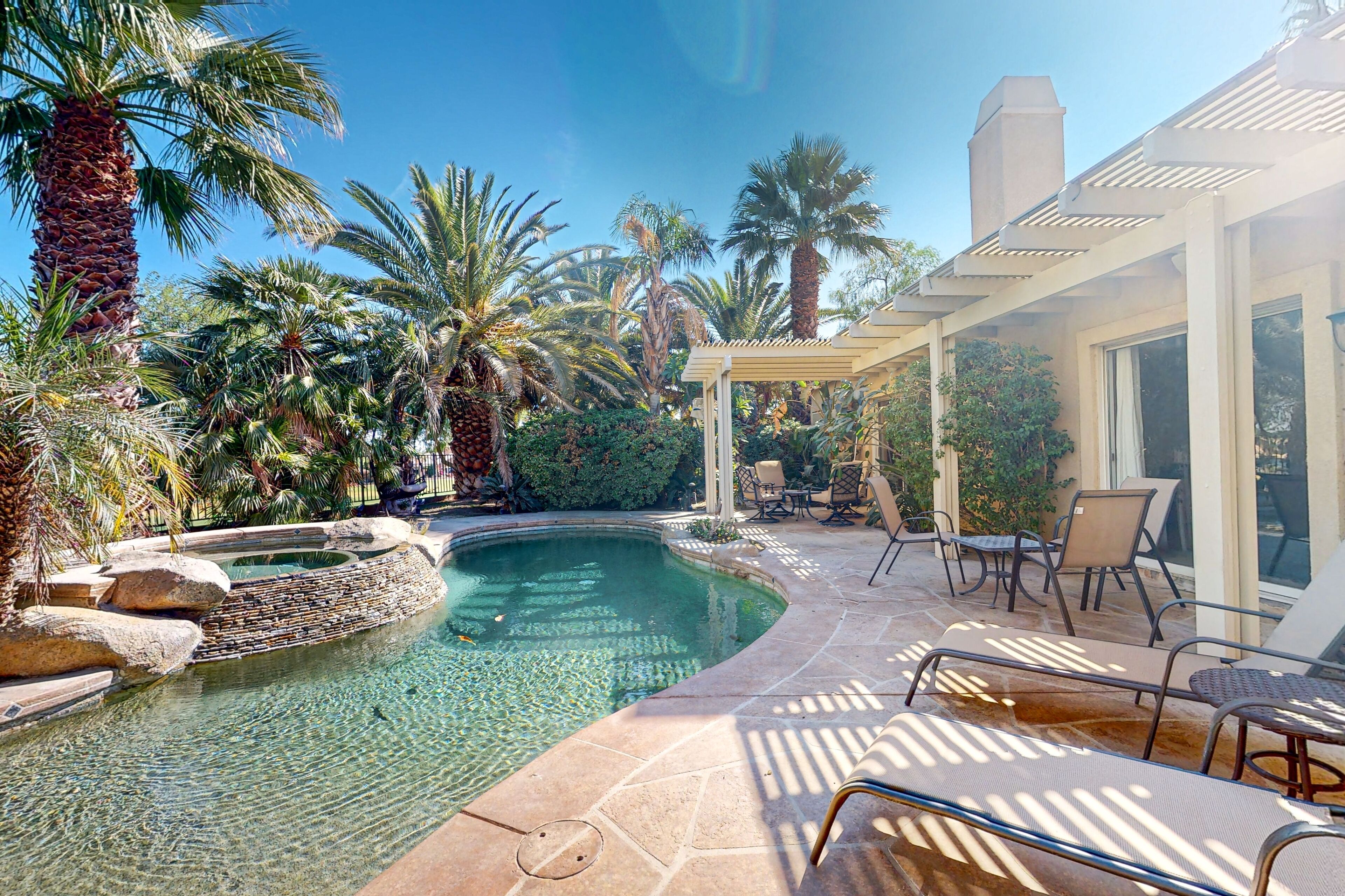 A swimming pool with clear water, surrounded by palm trees and a patio area with chairs and a pergola.