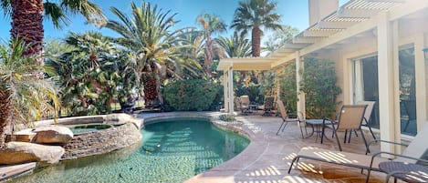 A swimming pool with clear water, surrounded by palm trees and a patio area with chairs and a pergola.
