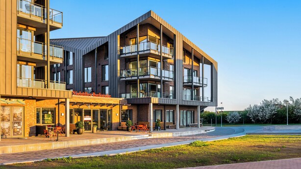 A modern multi-story building with a cafe on the ground floor, outdoor seating, and a landscaped area. - The View B04 Briese - Garden furniture, elevator, shared Sauna, parking space (Dagebüll)