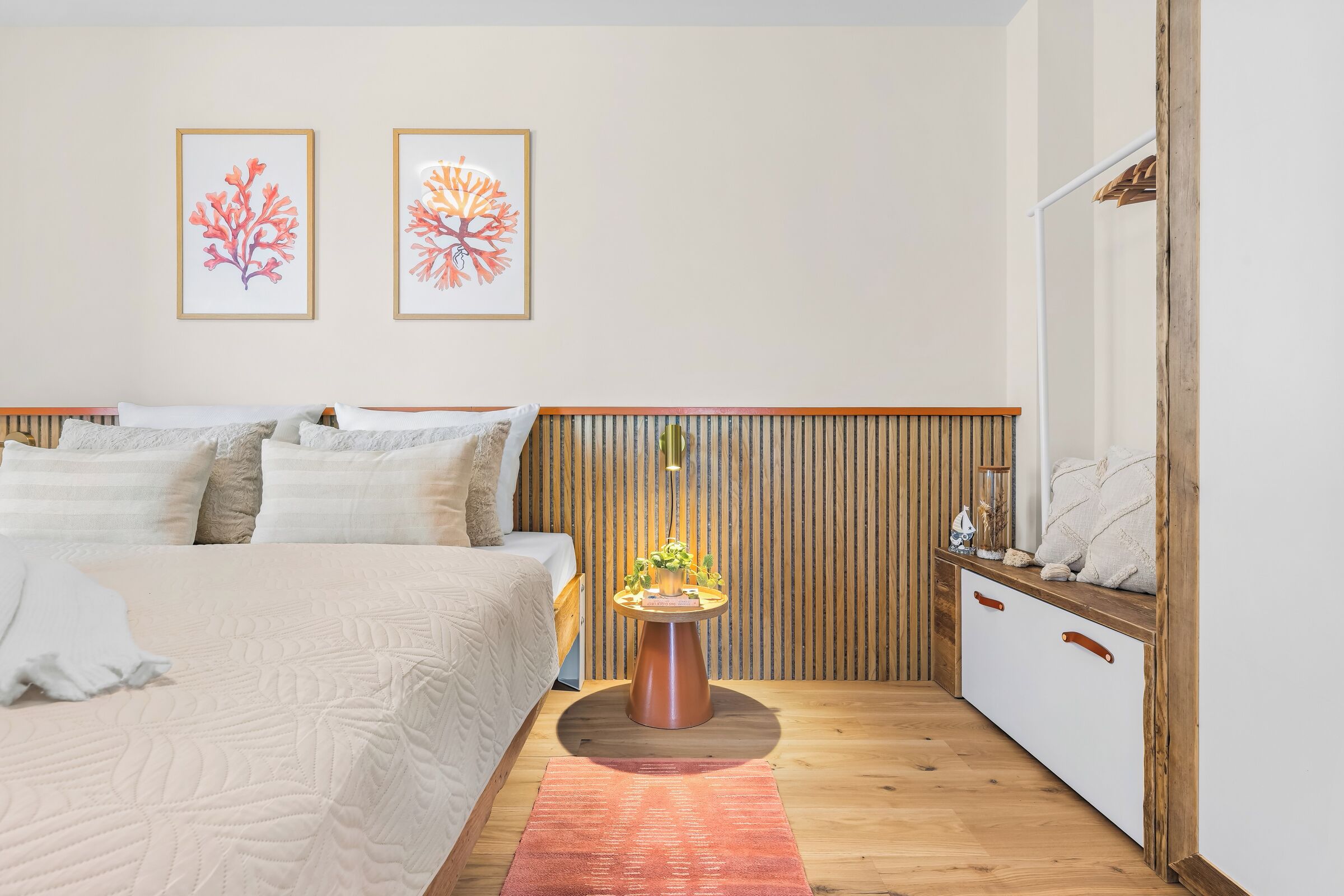 A bedroom with a bed, bedside tables, a nightstand, a wooden headboard, a wall-mounted shelf, and two framed artworks.