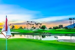 A golf course with a flag, palm trees, and a body of water reflecting the sky. - Glamorous home w/ mountain views, private pool, & on-site golf - 3 BR - #067092 (La Quinta)