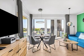 A modern living room with a flat-screen TV, a wooden cabinet, a dining table with chairs, a blue sofa, and a view through large windows. - The View B12 Zuhause bei Emi -Sea view,elevator, balcony, armchair, shared Sauna (Dagebüll)