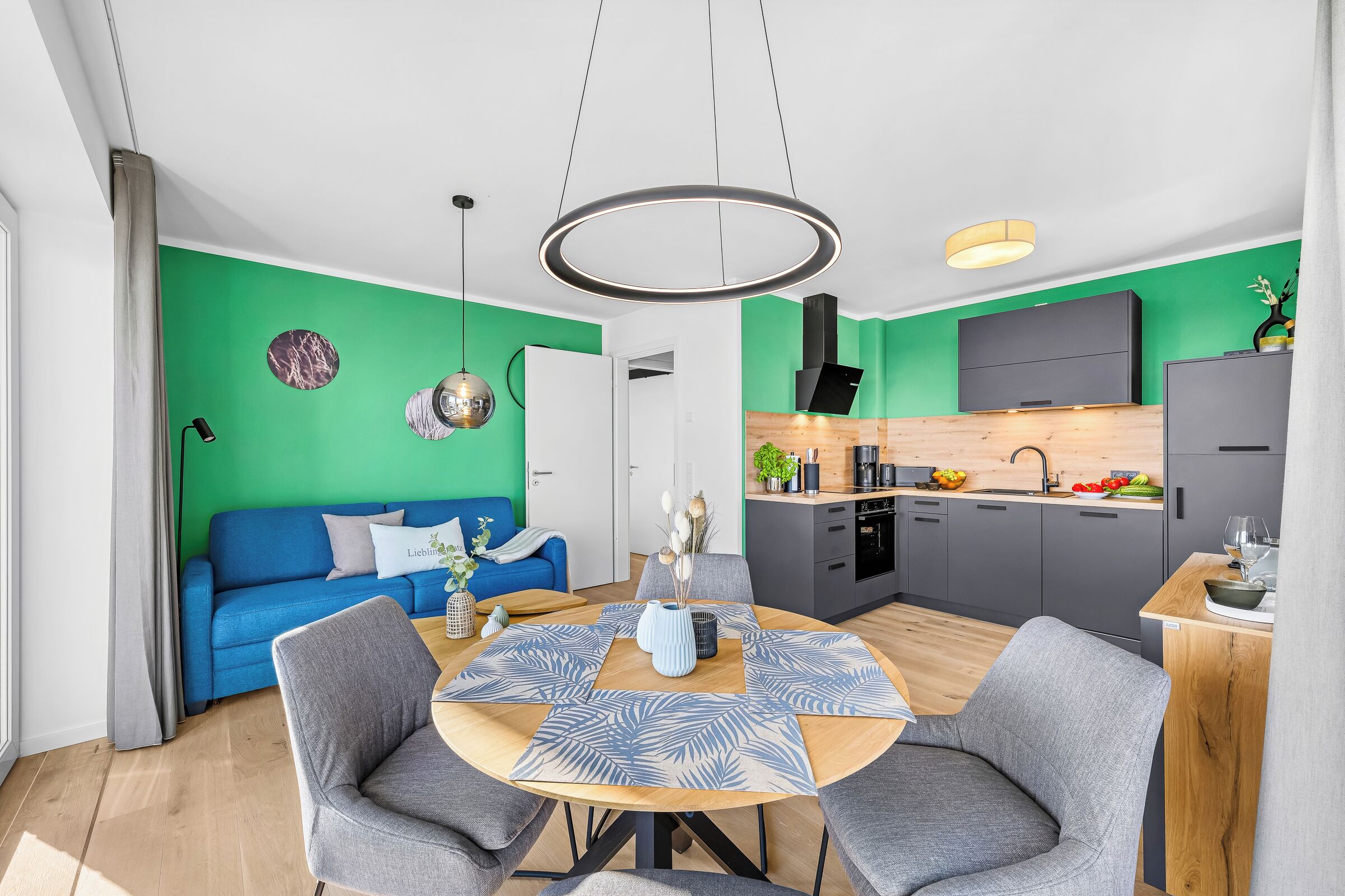 Ein moderner Wohnraum mit einem blauen Sofa, einem runden Esstisch mit grauen Stühlen, einer Küche mit einem schwarzen Dunstabzug und einer Akzentwand in Grün.