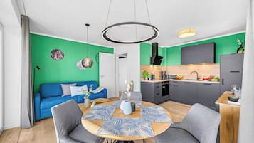 Ein moderner Wohnraum mit einem blauen Sofa, einem runden Esstisch mit grauen Stühlen, einer Küche mit einem schwarzen Dunstabzug und einer Akzentwand in Grün.