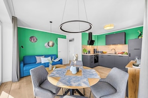 Ein moderner Wohnraum mit einem blauen Sofa, einem runden Esstisch mit grauen Stühlen, einer Küche mit einem schwarzen Dunstabzug und einer Akzentwand in Grün.