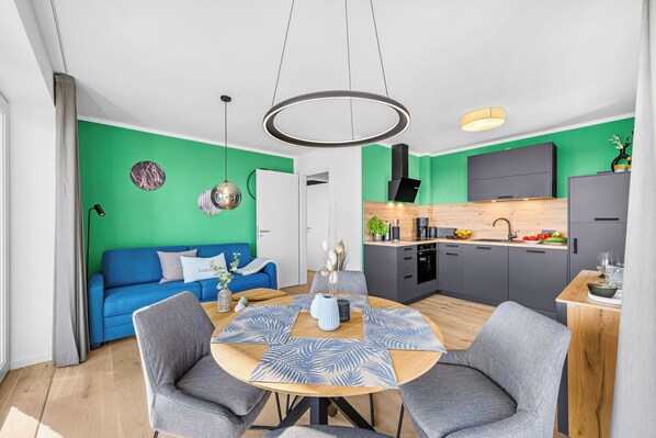 Un espacio habitable moderno con un sofá azul, una mesa de comedor redonda con sillas grises, una cocina con campana extractora negra y una pared de acento verde.