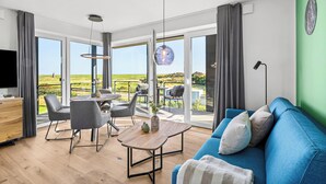 A modern living room with a blue sofa, a dining table with chairs, and a large window offering a view of a green landscape. - The View B12 Zuhause bei Emi -Sea view,elevator, balcony, armchair, shared Sauna (Dagebüll)