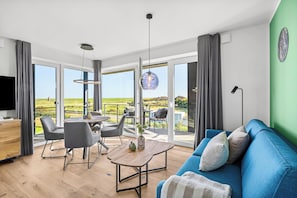 Ein modernes Wohnzimmer mit einem blauen Sofa, einem Esstisch mit Stühlen und einem großen Fenster, das einen Blick auf eine grüne Landschaft bietet.