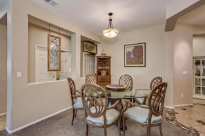A dining room with a glass table, six chairs, a chandelier, a wooden cabinet with glassware, and framed artwork on the walls. - Classic, dog-friendly home near golf with heated pool, enclosed yard & fireplace (Indio)