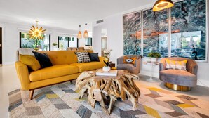 A modern living room with a yellow sofa, a round coffee table with a natural wood base, and a gray armchair. There are pendant lights, a large abstract painting, and a view of the outdoors through the windows. - Mtn-view home with private pool, large patio, streaming, fireplace, grill, & W/D (Palm Springs)
