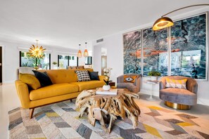 Ein modernes Wohnzimmer mit einem gelben Sofa, einem runden Couchtisch mit natürlicher Holzbasis und einem grauen Sessel. Es gibt Pendelleuchten, ein großes abstraktes Gemälde und einen Blick ins Freie durch die Fenster.