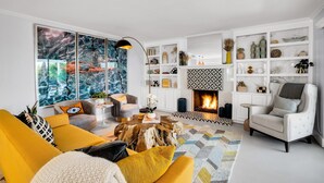 A modern living room with a fireplace, a large window with a cityscape view, and a bookshelf filled with decorative items. - Mtn-view home with private pool, large patio, streaming, fireplace, grill, & W/D (Palm Springs)